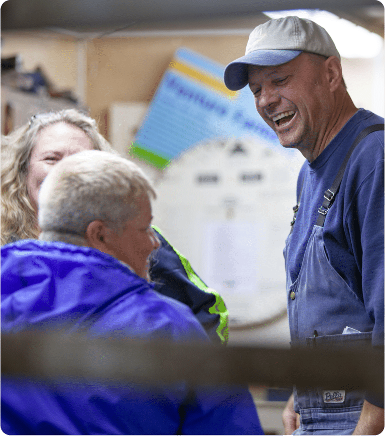 A farmer wearing a white and blue hat, blue shirt and blue overalls smiles and laughs with his team members in the office of their dairy farm in Upstate New York