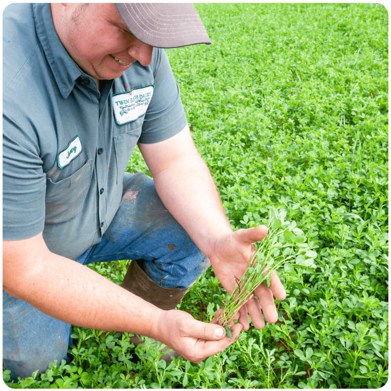 A farmer in a green shirt, blue jeans and brown rubber boots proudly smiles and displays a crop crown on his fields